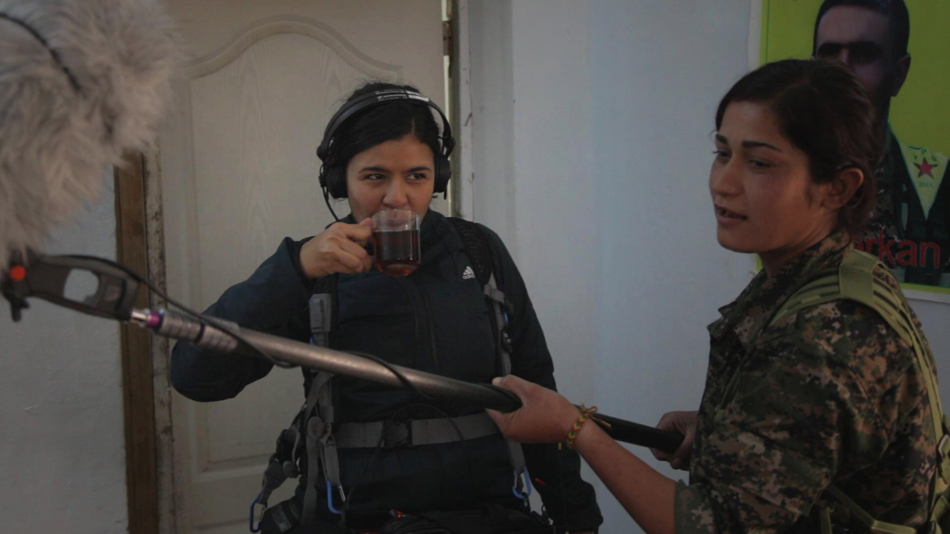 Ana wears her headphones and drink tea as a YPJ female soldier holds her boom mic.