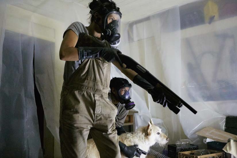 Donned with gas masks in a room covered in plastic, two people with weapons and a dog anxiously look for their target