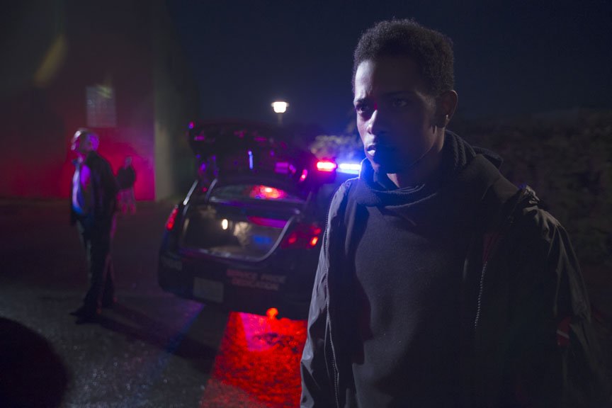 The character 'L' looks intently onward with police cars behind him.