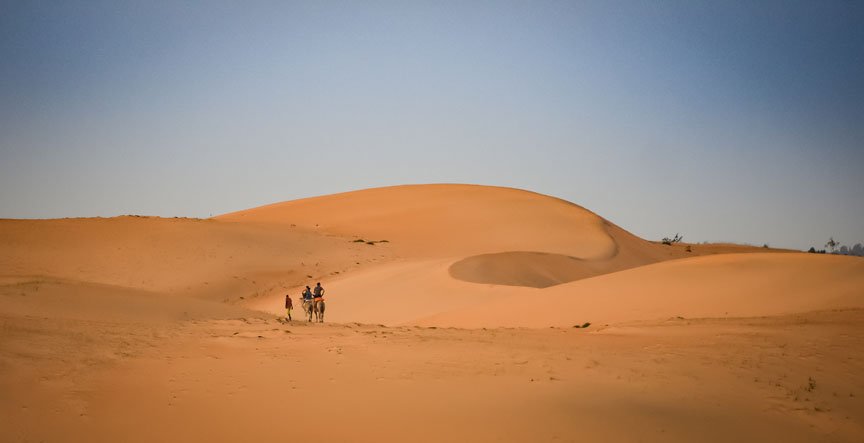 Two people ride donkeys through the completely sand-covered desert hills.