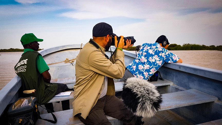 Three men sit in a boat while one man records and another photographs.