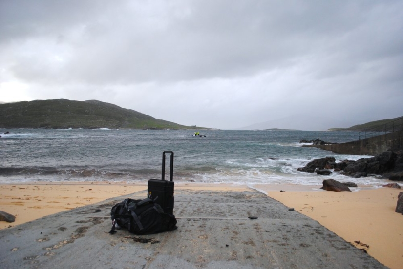 A bag and suitcase lay in the sand as a boat approaches the shoreline.