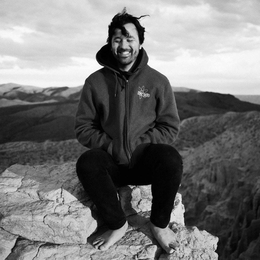 A man sits barefoot on a rock with mountains behind him.