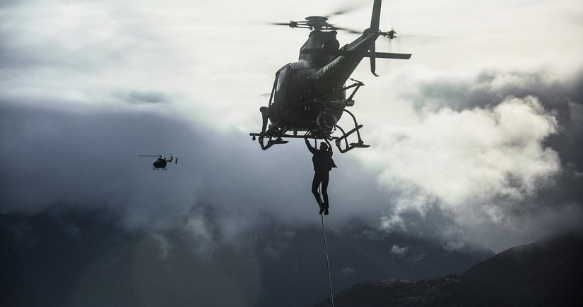 Tom Cruise hangs from a helicopter over a cloudy moutain range.