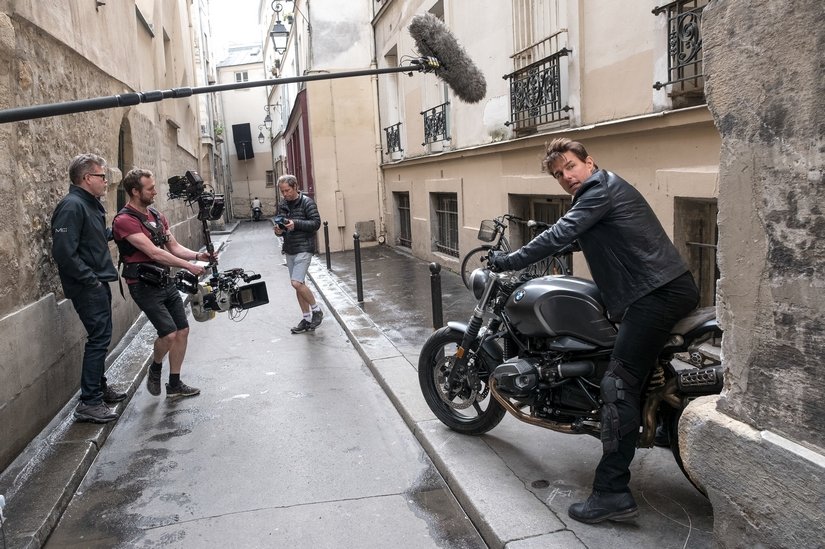 A boom mic extends to Tom Cruise who sits on a BMW motorcycle in a European alley.