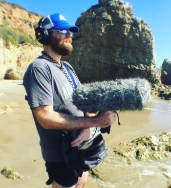 A man with a blonde beard records ambience on a shoreline.