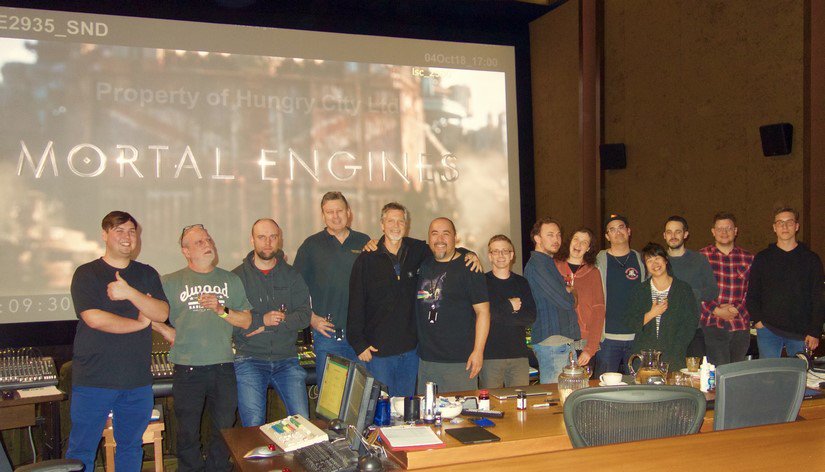 14 men and 2 women stand in a screening room with the title Mortal Engines on the projector behind.