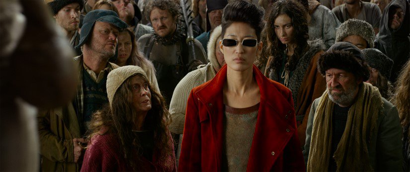 A woman with a pompadour, red coat, and square sunglasses stands tall in a crowd of townspeople.