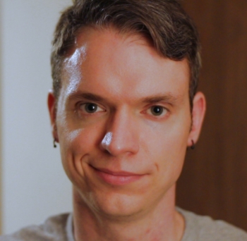 A man with short hair and earrings smiles slightly wryly.