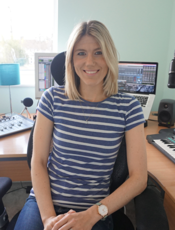 A woman with blonde hair smiles as she sits at her workspace.