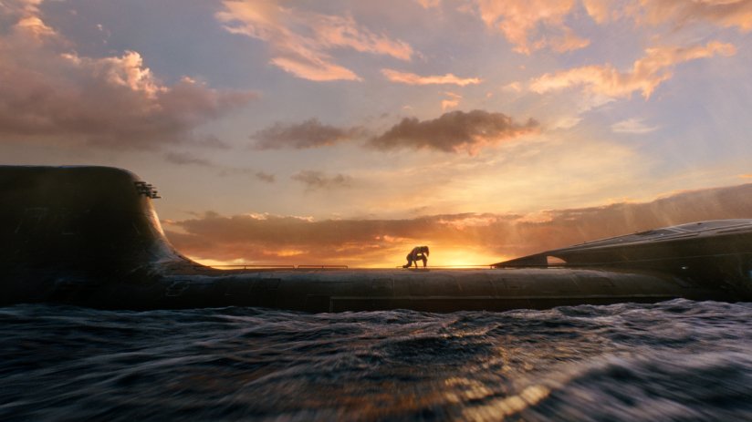 Aquaman kneels next to a shore at sunrise.