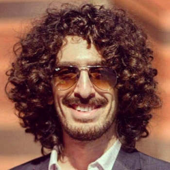 A man with curly brown hair and dark sunglasses smiles.