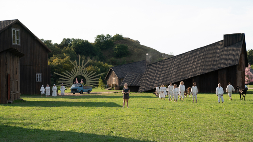 Wooden farm building line an open field full of people dressed in white.